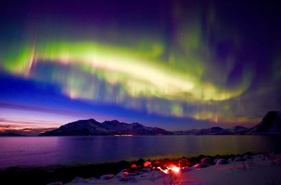 Colorful Northern Lights over the fjord and a island, on blue and sunset sky. Campfire on the beach.