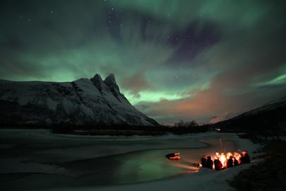 Guests around the camp fore at the frozen river while the Northern lights decorate the sky