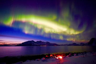 Camp fire by the fjord with colorful Aurora over the ocean with sunset glow sky