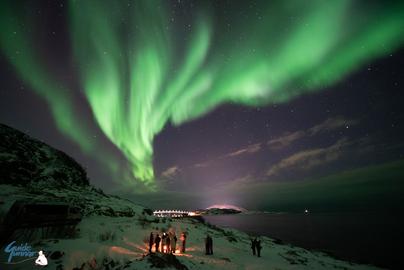 People around the campfire by the fjord and the bridge with Northern Lights on the sky