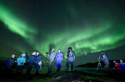 Many people with camera and tripods under the Northern Lights, by the fjord.